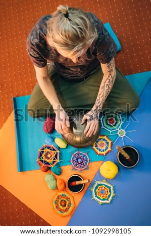Young blond caucasian man with henna tattoo on his hand playing hang drum with fingers sitting on mat in class in front of yarn, knitted mandalas. Top view.