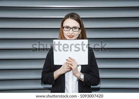 A young woman in a business suit is near the office. Business woman in glasses and a notebook. The girl is making notes on her daily party. Good girl smiles. The lady stands near a gray wall.