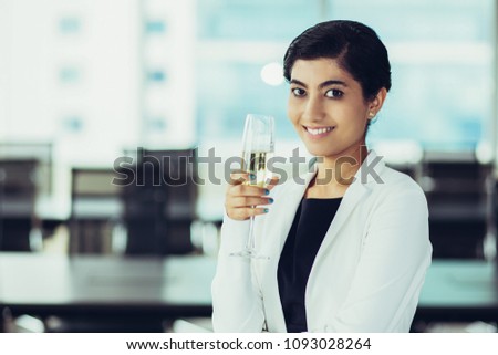 Portrait of successful young Asian businesswoman wearing suit standing in office holding champagne flute, looking at camera and smiling