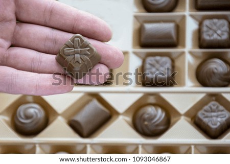 Focus on human arm holding sweet that has form of present box with decorative bow and ribbons. Carton with confection collection stored into special shapes cells.