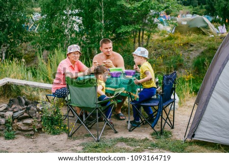 children with grandmother and father sit at a folding table on a picnic with a tent in the summer