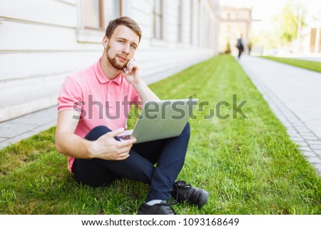 Handsome man sitting on the grass in the city with a laptop and talking on the phone, job search, rest on the Internet