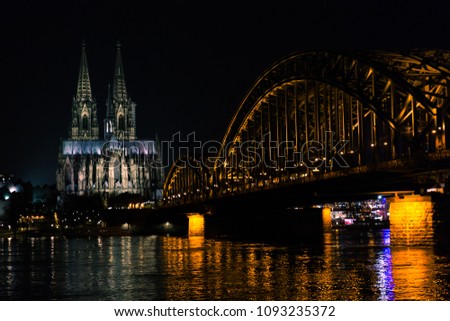 Rhine with flood in the evening. In the background the Cologne Cathedral. Right in the picture the illuminated Hohenzollern bridge. Glittering reflections on the water.