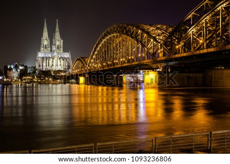 Rhine with flood in the evening. In the background the Cologne Cathedral. Right in the picture the illuminated Hohenzollern bridge. Glittering reflections on the water. Long exposure.
