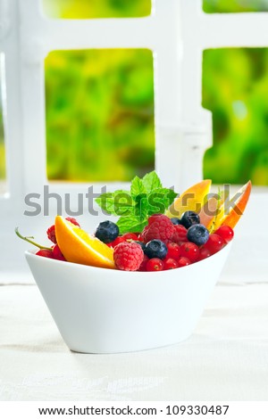 Bowl of fresh berries including raspberries, blueberries and redcurrants, garnished with sliced oranges and mint on an indoor table with copyspace