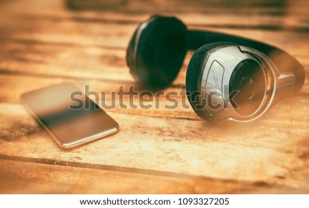 A 45 degree view of a set of wireless headphones and mobile phone lying on a rustic wooden table outside. Styling and grain effect added to image.