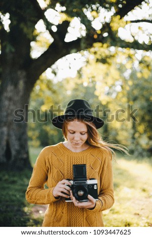 Young curious white beautiful woman in warm yellow sweater with ginger hair in a wool hat stay in a wild woods taking picture with soviet vintage film camera looking through viewfinder