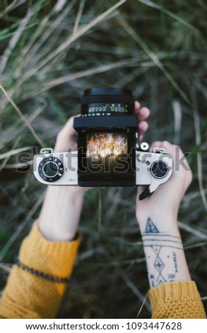 Young white girl in a ginger knitted sweater with tattoed hand holding vintage soviet medium camera and taking a picture of warm landscape