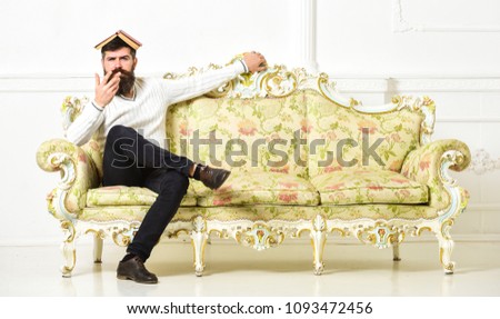 Macho sits with open book on head, like roof. Overstudy concept. Man with beard and mustache sits on sofa, white wall background. Guy overdid with studying, fed up of reading old boring book