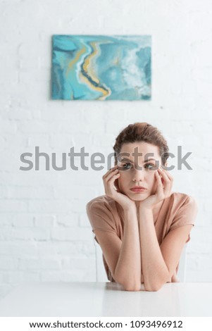 thoughtful young woman sitting at table in front of white brick wall with picture