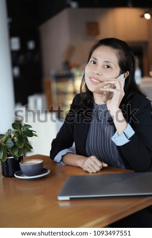 Smiling asian business woman using phone in coffee shop, laptop computer on the wooden table.