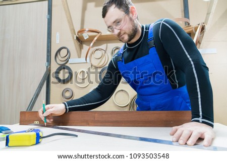 A bearded tired handsome carpenter on a table with a pencil draws a sign on the board. At the workplace there is an ordinary master dressed in blue cambenizon with a cruel haircut and saved glasses