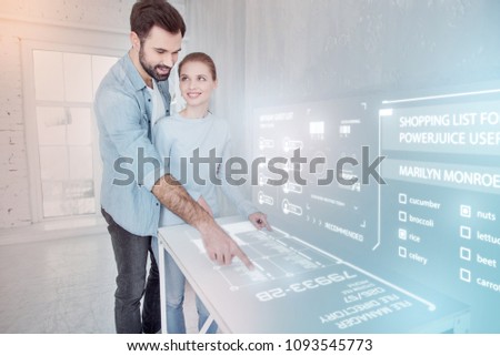 Pleasant activity. Cheerful young web designer feeling happy while standing near his smiling girlfriend and touching the screen of his device
