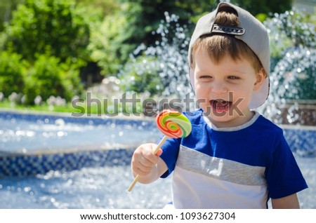 happy boy of two years is sitting near the fountain in the summer and eats a delicious multi-colored candy on a stick. A hot, sunny summer. Outdoor recreation, snack with street food