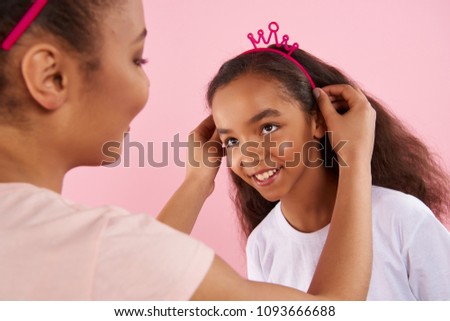 African American mother puts crown on head of small daughter. Isolated on pink background. Studio portrait.
