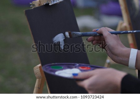 close up girl paints a painting on the canvas with the help of paints. A wooden easel keeps the picture. Summer is a sunny day, sunset