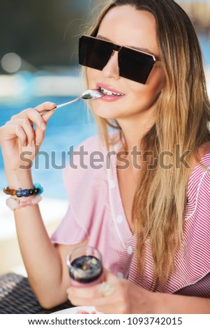 Outdoors lifestyle fashion image of young charming girl having breakfast. Holding in her hand glass of pudding, small spoon. Eating and enjoying vacation. Wearing stylish sunglasses, summer dress