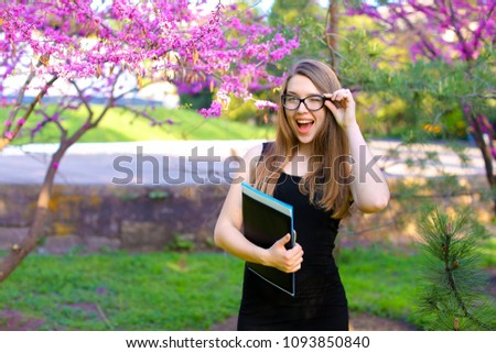 Female student touching glasses and standing in park with black folder near trees in blossom. Concept of education, positive emotions and spring.