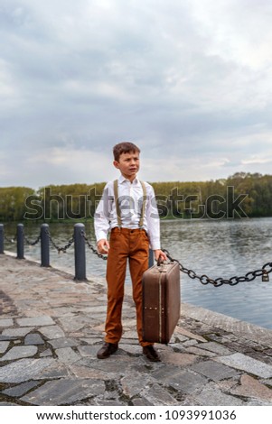 little boy simpleton in an old-fashioned rustic clothes with a vintage suitcase standing at the arrival station on the pier of the lake river Royalty-Free Stock Photo #1093991036