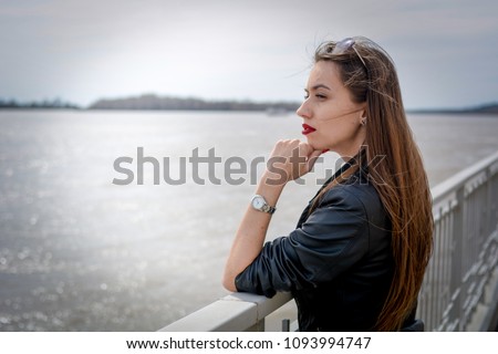 Fashionable beautiful woman in a black jacket on a summer day against the background of the city river Royalty-Free Stock Photo #1093994747