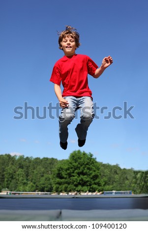 Joyful boy dressed in red T-shirt jumps on trampoline at sunny summer day