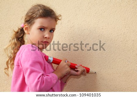Girl paints by big red pencil on yellow wall and looks into camera.