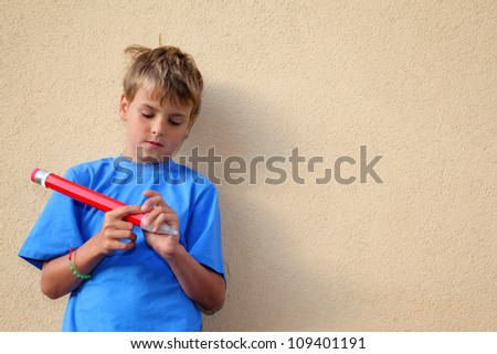 Boy with big red pencil stands near yellow wall and closes pencil by cap.