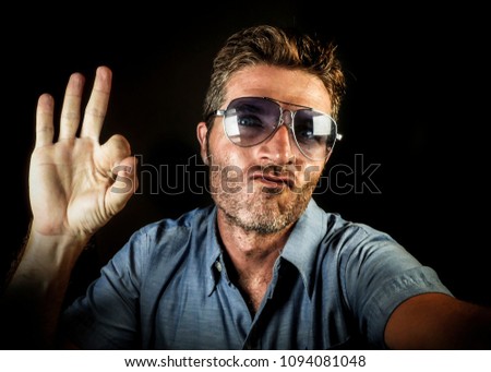 crazy happy and funny guy with sunglasses and modern hipster look taking selfie self portrait picture with mobile phone camera smiling cool and giving ok sign with fingers posing as a winner