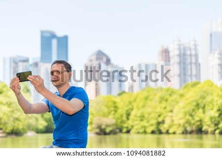 Young man sitting taking selfie in Piedmont Park in Atlanta, Georgia with scenic water, and cityscape skyline of urban city skyscrapers downtown, Lake Clara Meer