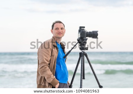 Young man professional photographer taking pictures and video of beach sunset in Florida panhandle, with jacket, wind, beach waves, tripod