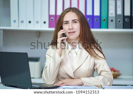 Portrait of a beautiful brunette girl manager working in the office behind a laptop and talking on a smartphone. She sits right in front of the camera and looks serious.