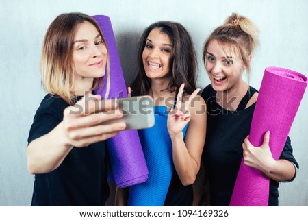 three attractive and young female girlfriends hold a roll yoga mat and do selfie on the phone. concept of yoga practice and female friendship