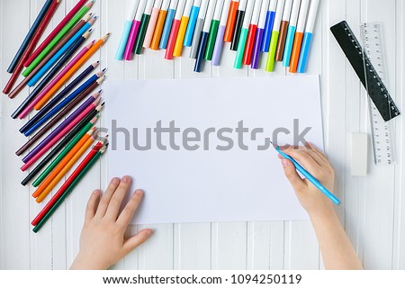 The child's hands are painted with colored pencils on a white sheet of paper on a wooden table.