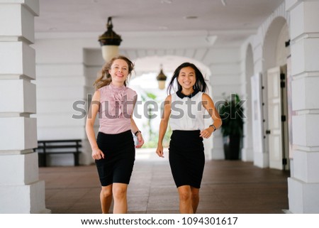 A Chinese woman and Caucasian white woman walk down a corridor today. They are both dressed in office attire and the diverse pair are talking as they walk.
