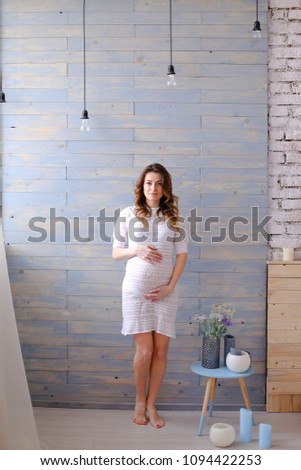 Pregnant woman standing in white dress, wooden wall and lamps in background. Concept of pregnance and interior.