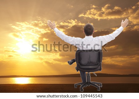 Man with raised hands sitting on chair on asphalt road on orange sunset