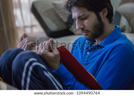 Close-up shot of a young man sitting on the floor leaning his back on the couch reading a red book.