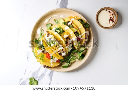Variety of vegetarian corn tacos with vegetables, green salad, chili pepper served on ceramic plate with cream sauce over white marble background. Top view, copy space.