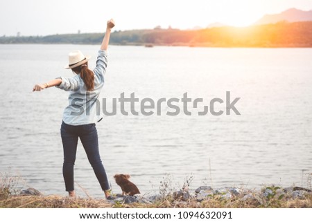 Happy asian woman stands and stretching arm for relaxing in nature outdoor sunset beside river with dog. Young traveler enjoying with pet in summer vacation. Holiday, relaxation and freedom concept.