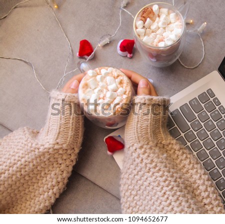 Hot chocolate with marshmallow in a woman hand , with Laptop and  tiny Santana Claus hats in the background.