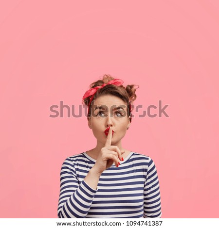 Portrait of pinup girl wears clothes in retro style, shows silence sign and looks secretly upwards, tells secret information, poses against pink background with blank copy space for your text