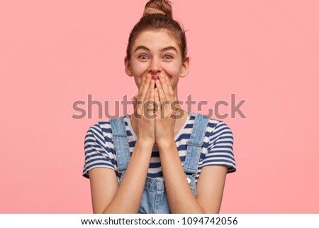 Beautiful female giggles joyfully, covers mouth with palms, glad to recieve letter of commendation for diligent work, has hair bun, wears striped casual t shirt with jean overalls, being glad
