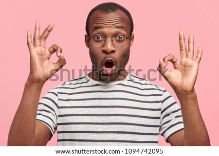 Emotional surprised dark skinned male shows ok sign with both hands, demonstrates that everything is fine, shows his approval, gestures against pink wall. Body language and facial expressions