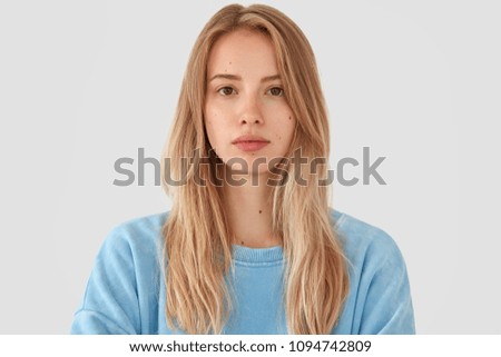 Headshot of tender beautiful young female model with fair hair, wears blue top, has healthy pure skin, looks seriously at camera, poses against white concrete wall. Cute teenager feels confident