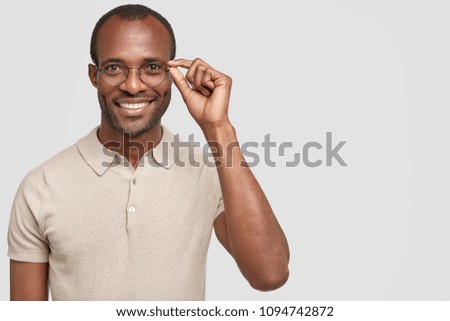 Positive African American male with shining smile, wears casual clothes, looks through spectacles, being satisfied with pleasant conversation, isolated on white background, enjoys positive news