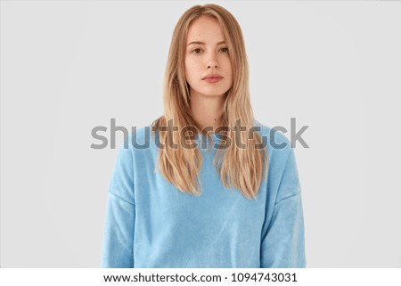 Waist up portrait of serious good looking blonde female in loose light blue casual sweater, looks attentively and pensively at camera, enjoys domestic atmosphere, isolated on white concrete wall