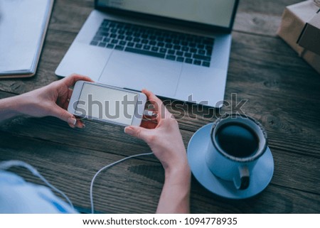 Cropped image of woman's hand holding modern smartphone while sending multimedia files to laptop computer editing before publish with mock up screen connected to free wireless internet in coffee shop.