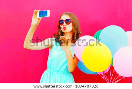 Woman taking selfie picture by smartphone blowing lips sending sweet air kiss over colorful balloons a pink background