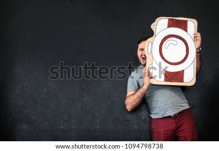 A man is a photographer with a large painted camera. Props. Funny Face. Dark gray background. Place for text. The photographer shoots at the camera. Picture of the camera. Focussed face. Profession. Royalty-Free Stock Photo #1094798738