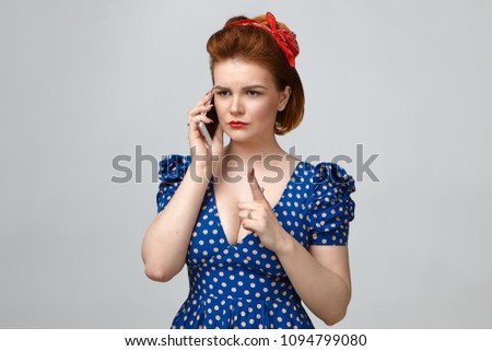 Picture of angry young mother in vintage outfit having strict look, raising index finger in front of her as warning sign, talking on smart phone with her naughty son who is still out with friends